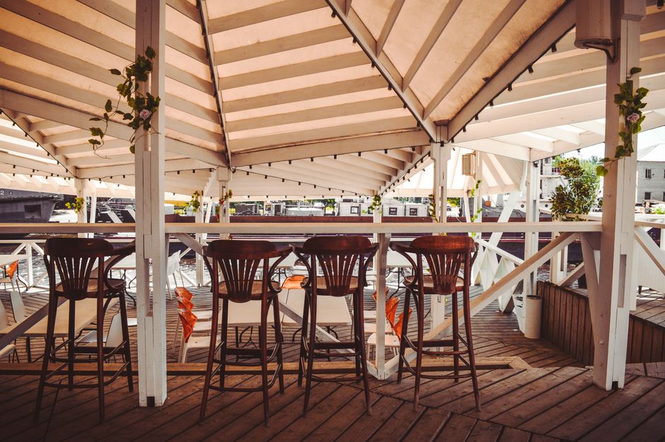Image of an outdoor seating area on a wooden deck beneath a large beige canopy with white framing, featuring five dark wooden bar stools with white cushions arranged around a table, demonstrating surf